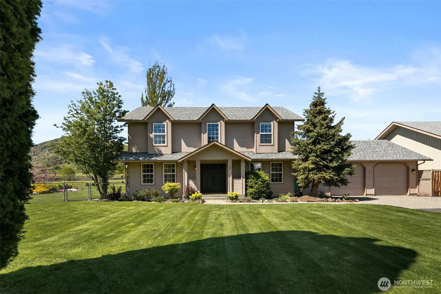Two-story tan house with gray roof, green lawn, and trees under a blue sky. A garage is attached to the right side of the house.