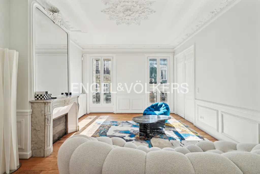 Bright living room with white walls, hardwood floors, and ornate ceiling. A marble fireplace, blue chair, and patterned rug add color.