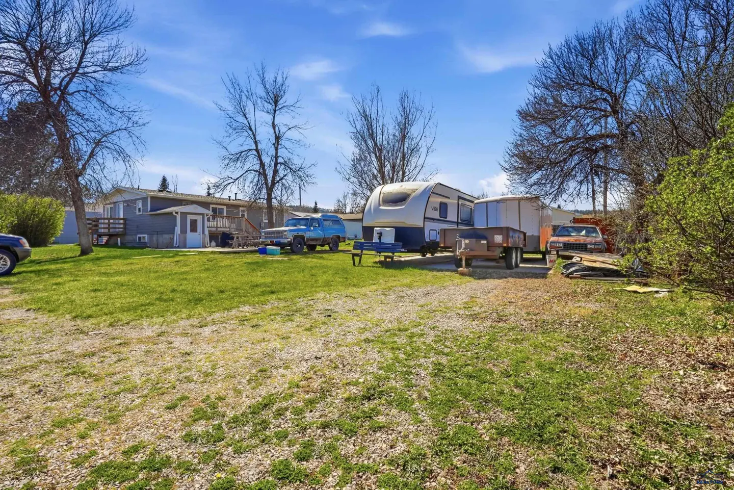 A wide shot of a property with a blue house, RV, blue truck, and trailers on a green lawn under a blue sky.