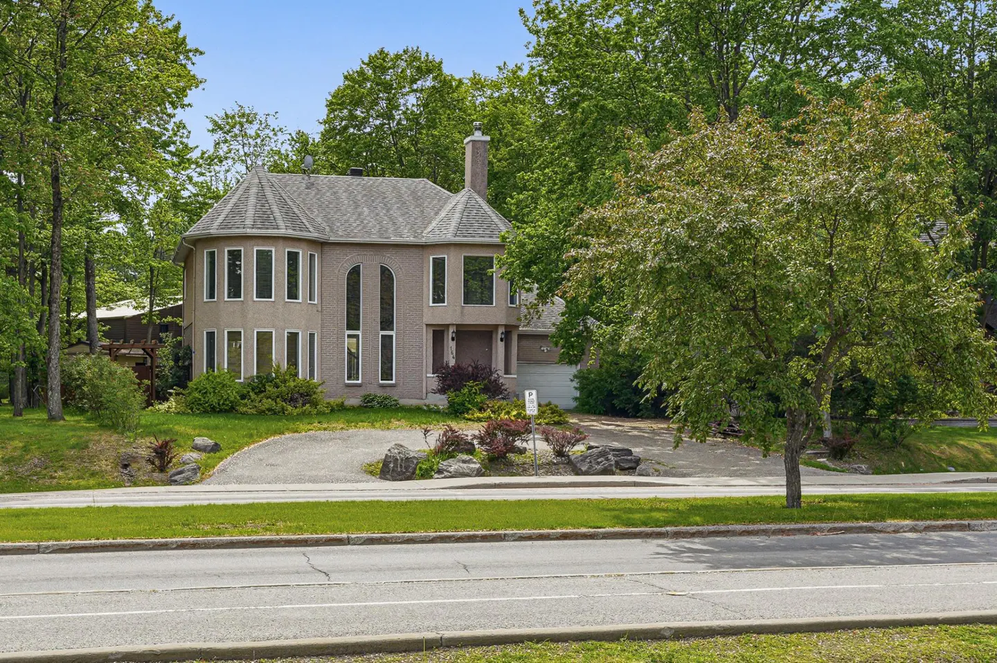Two-story tan brick house with a turret, arched windows, and a gravel driveway, surrounded by lush green trees.