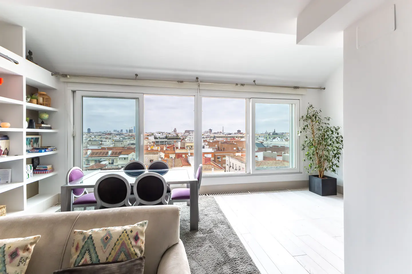 Bright, modern living room with a large window overlooking a city. A dining table with purple chairs sits near the window. A beige sofa is in the foreground.