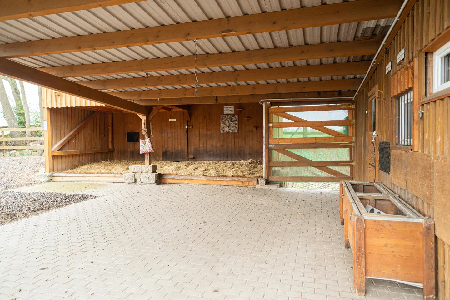 Wooden horse stable with hay and a metal roof. A wooden trough sits on a brick floor.