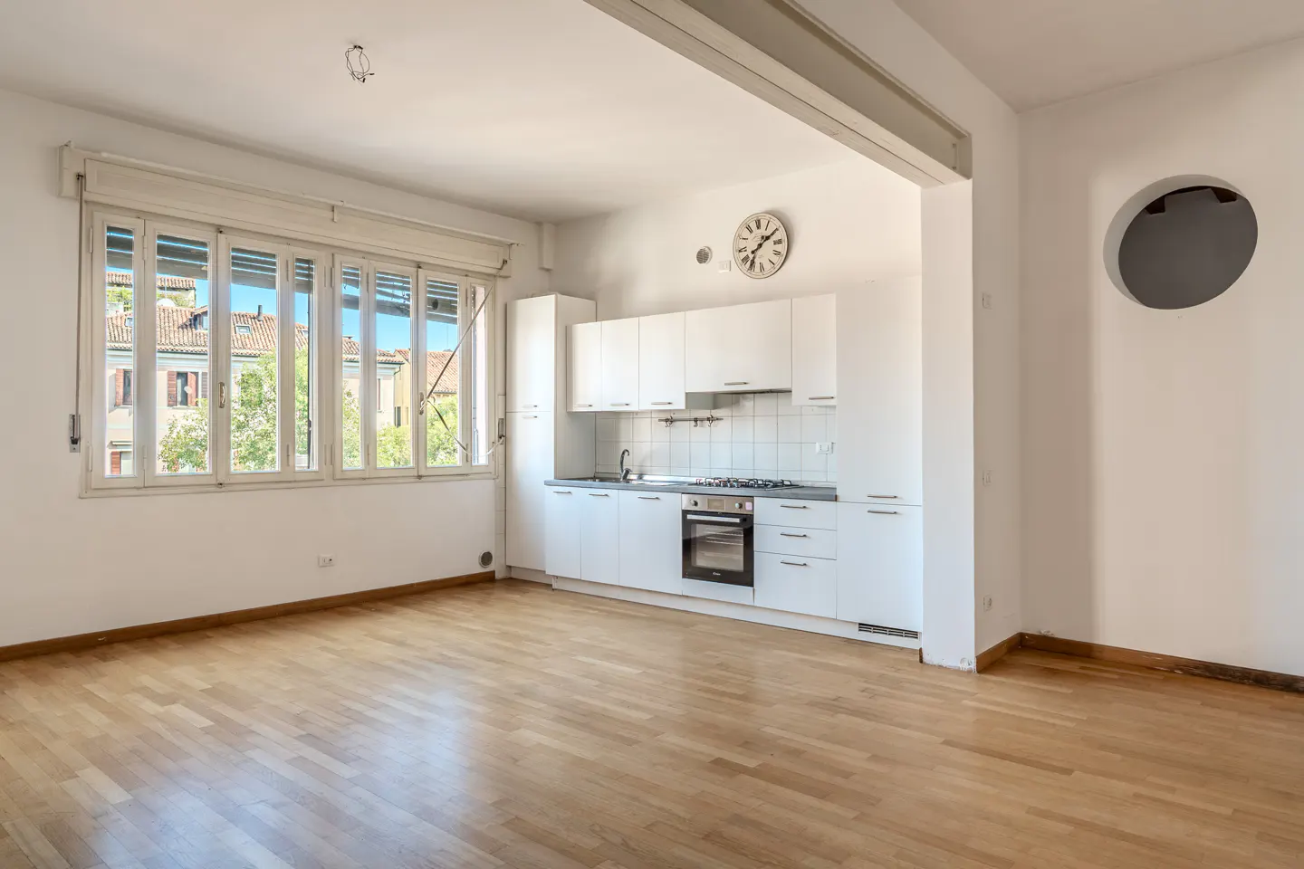 Bright, open-plan room with light wood floors, white kitchen cabinets, a large window, and a unique circular wall cutout.