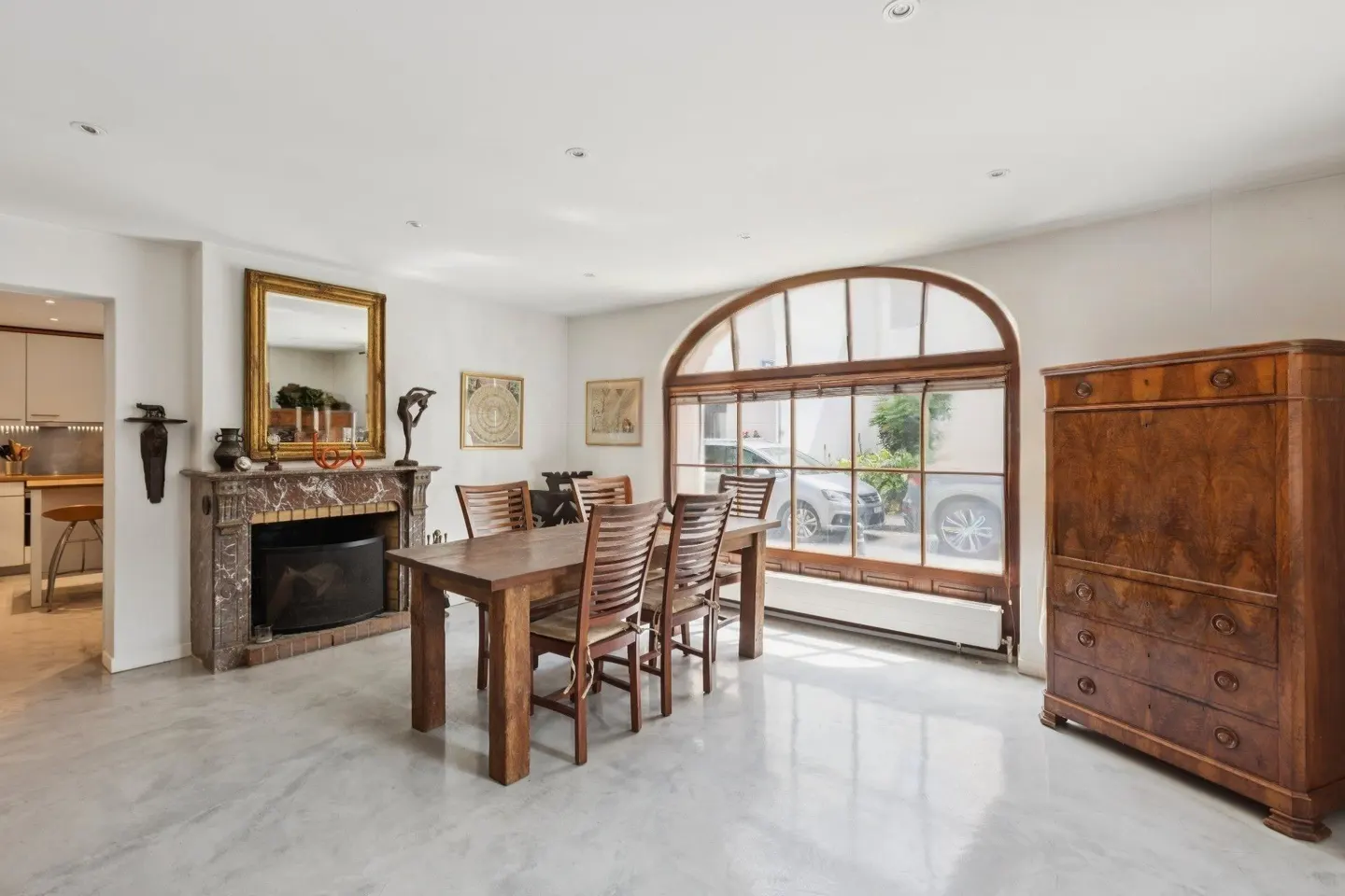 A dining room with a wooden table and chairs, a fireplace with a gold-framed mirror, and a large arched window.