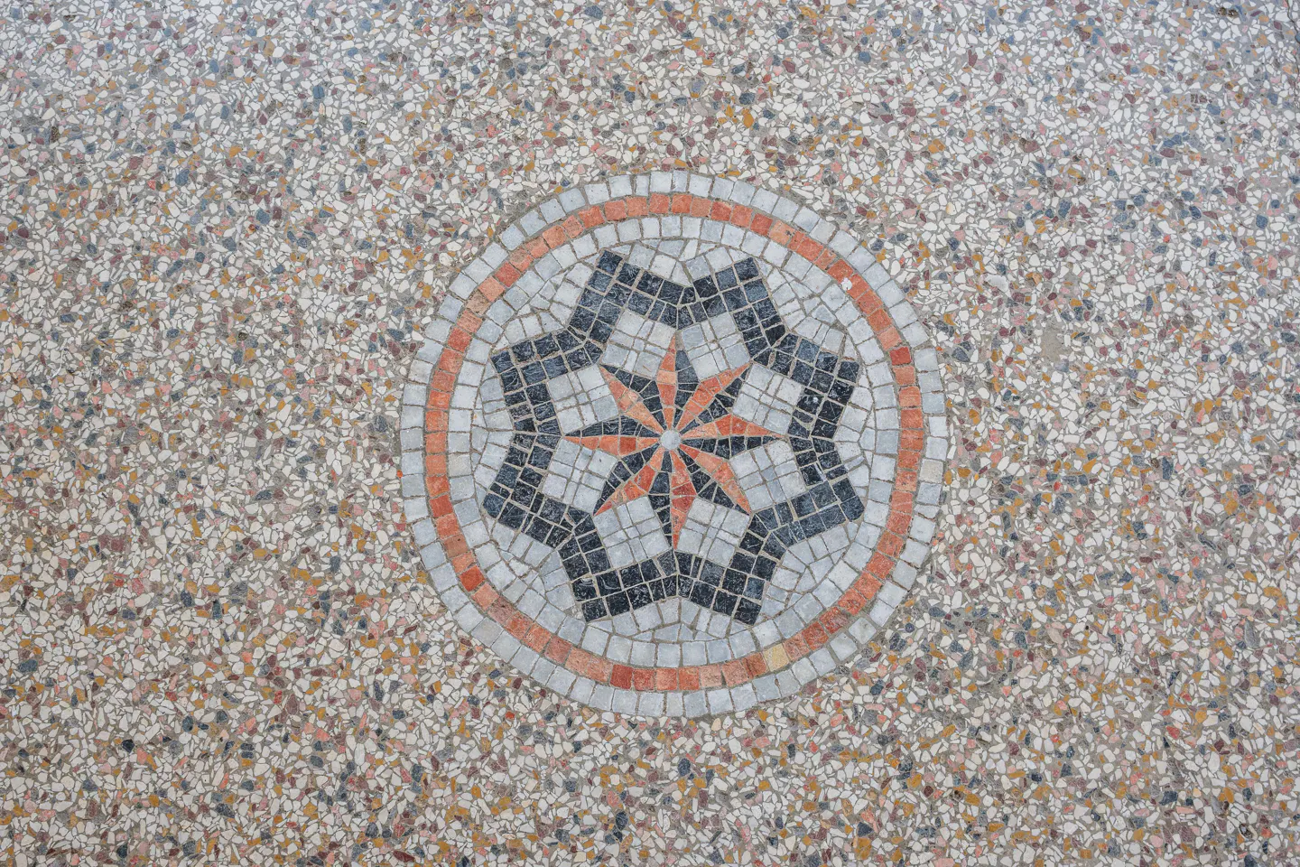 A circular mosaic medallion with a geometric star pattern in black, white, and orange, set in a speckled terrazzo floor.