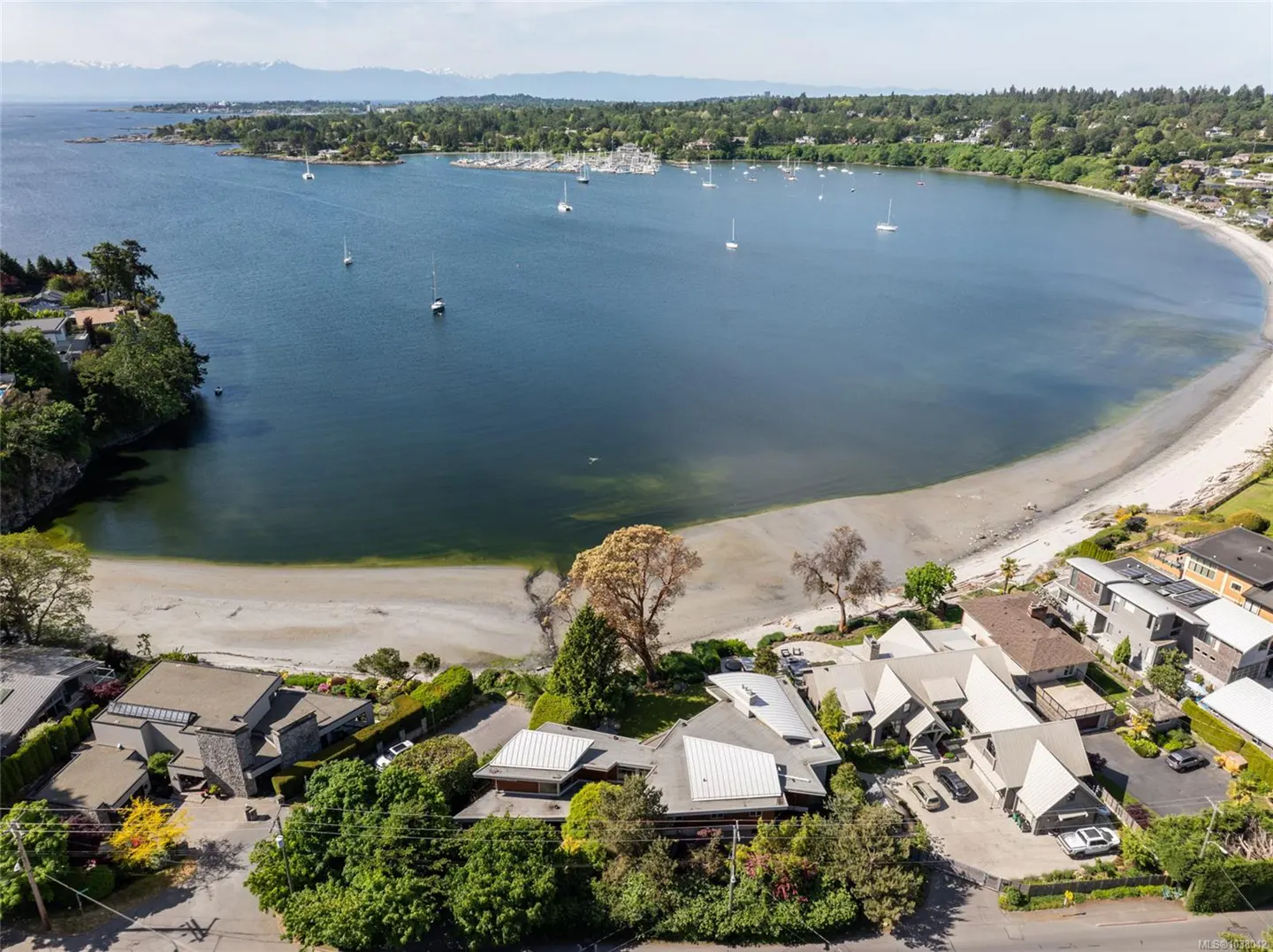 Aerial view of waterfront homes on a sandy beach, with sailboats on the blue water and mountains in the distance.
