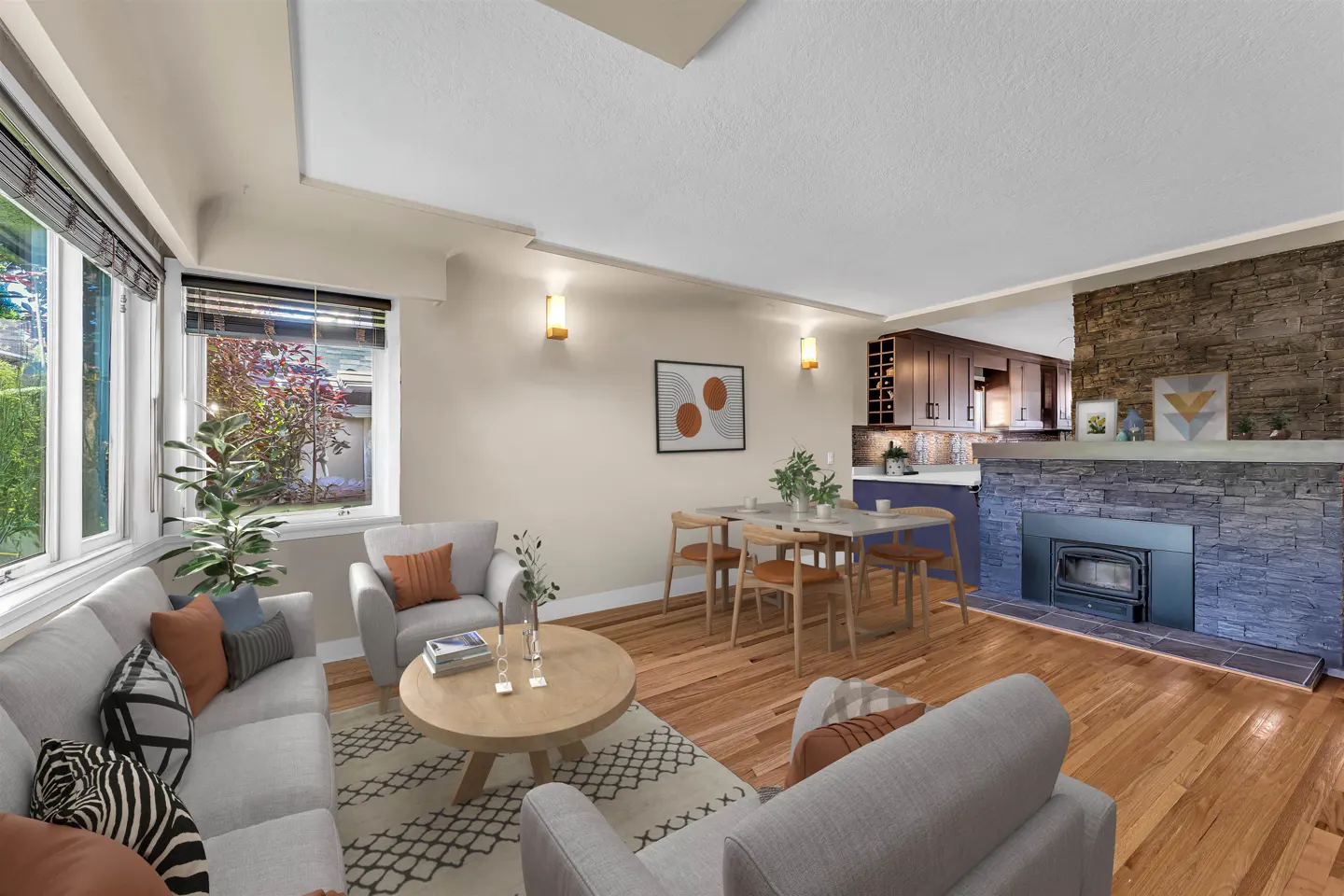 Living room with hardwood floors, a stone fireplace, and gray sofas. A dining table sits near an open kitchen. Natural light streams through large windows.