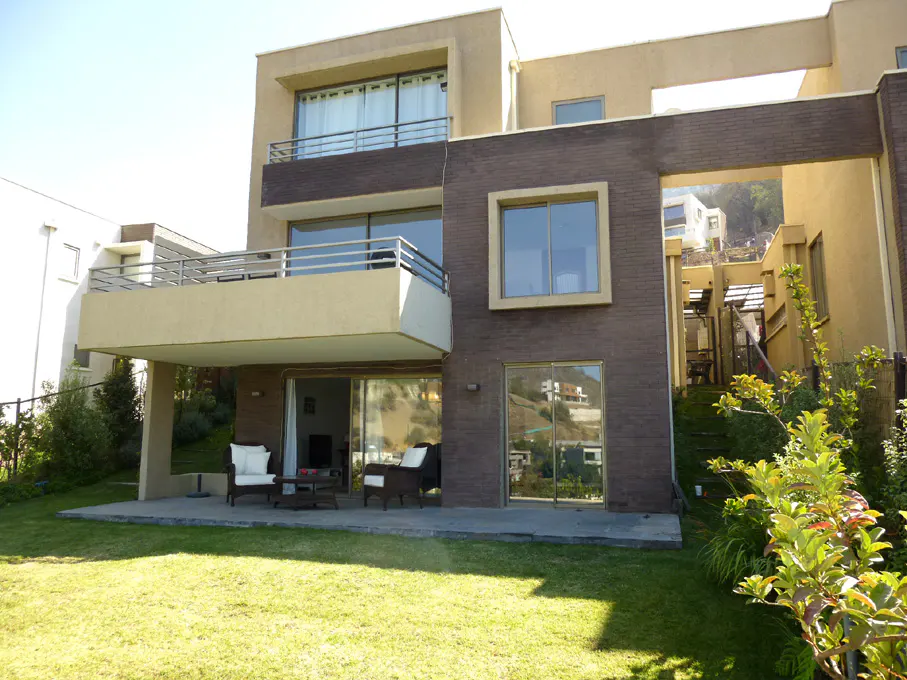 A modern, multi-story home with a lawn. The house is beige and brown with balconies and large windows. Patio furniture sits on a stone patio.