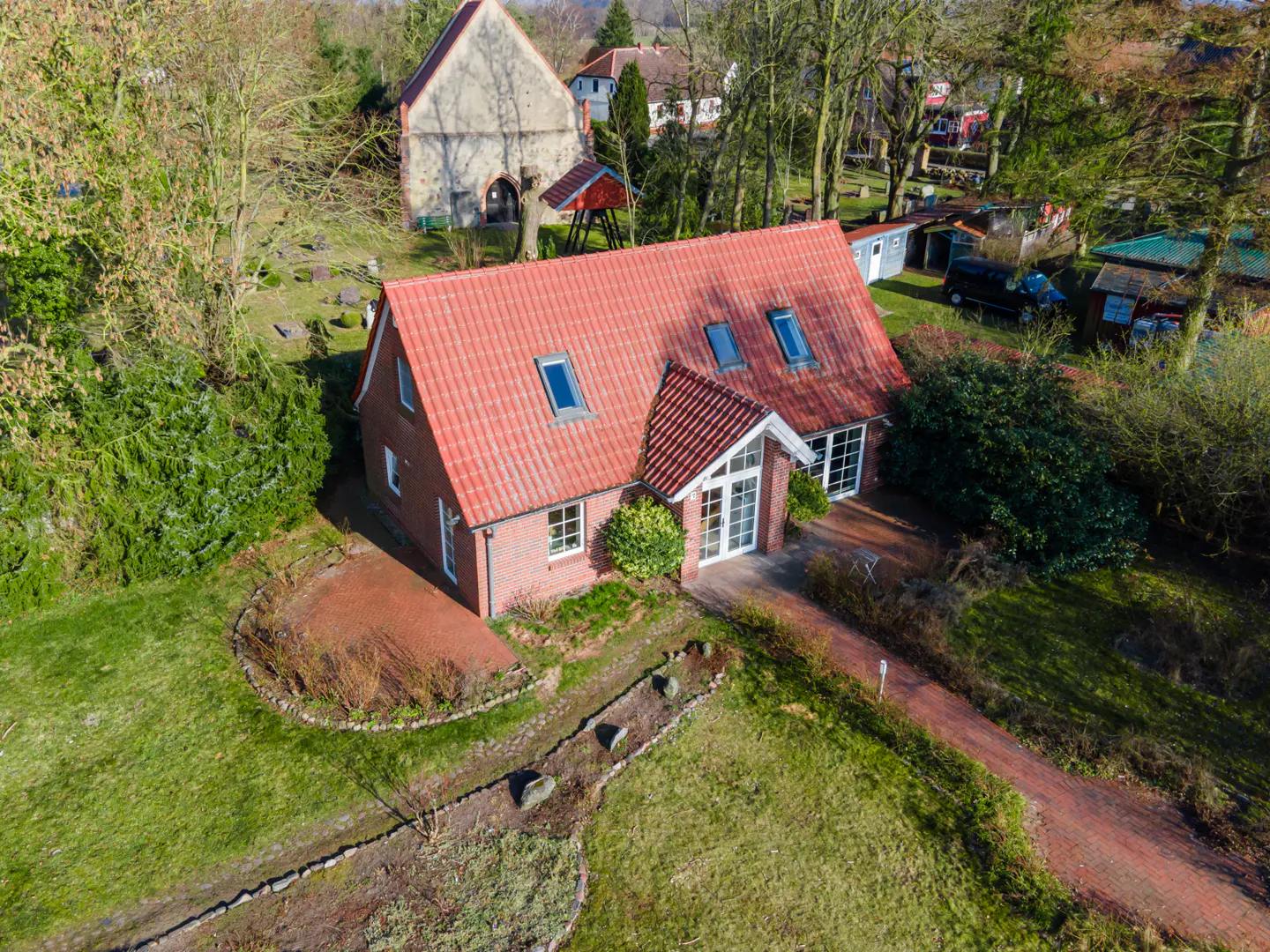 Aerial view of a red-roofed brick house with skylights, surrounded by green lawns and trees. A brick path leads to the front door.