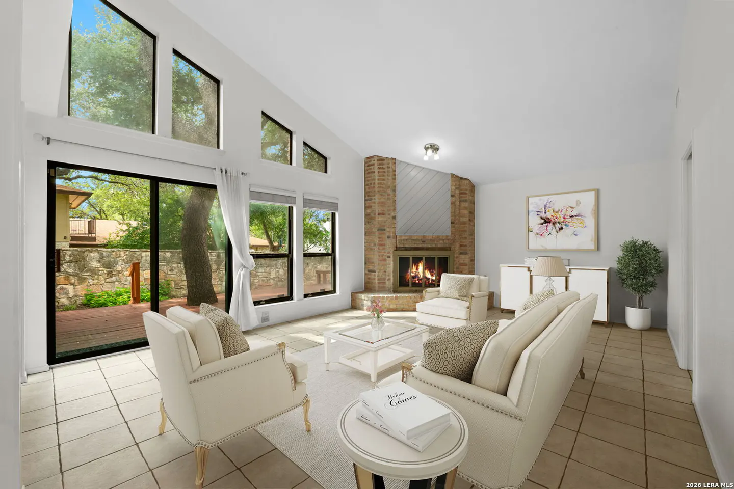 Bright living room with a brick fireplace, white furniture, and large windows overlooking a green yard.