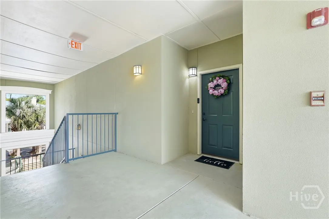 Hallway with blue railing, exit sign, and a blue door with a floral wreath. A black "hello" mat sits in front of the door.
