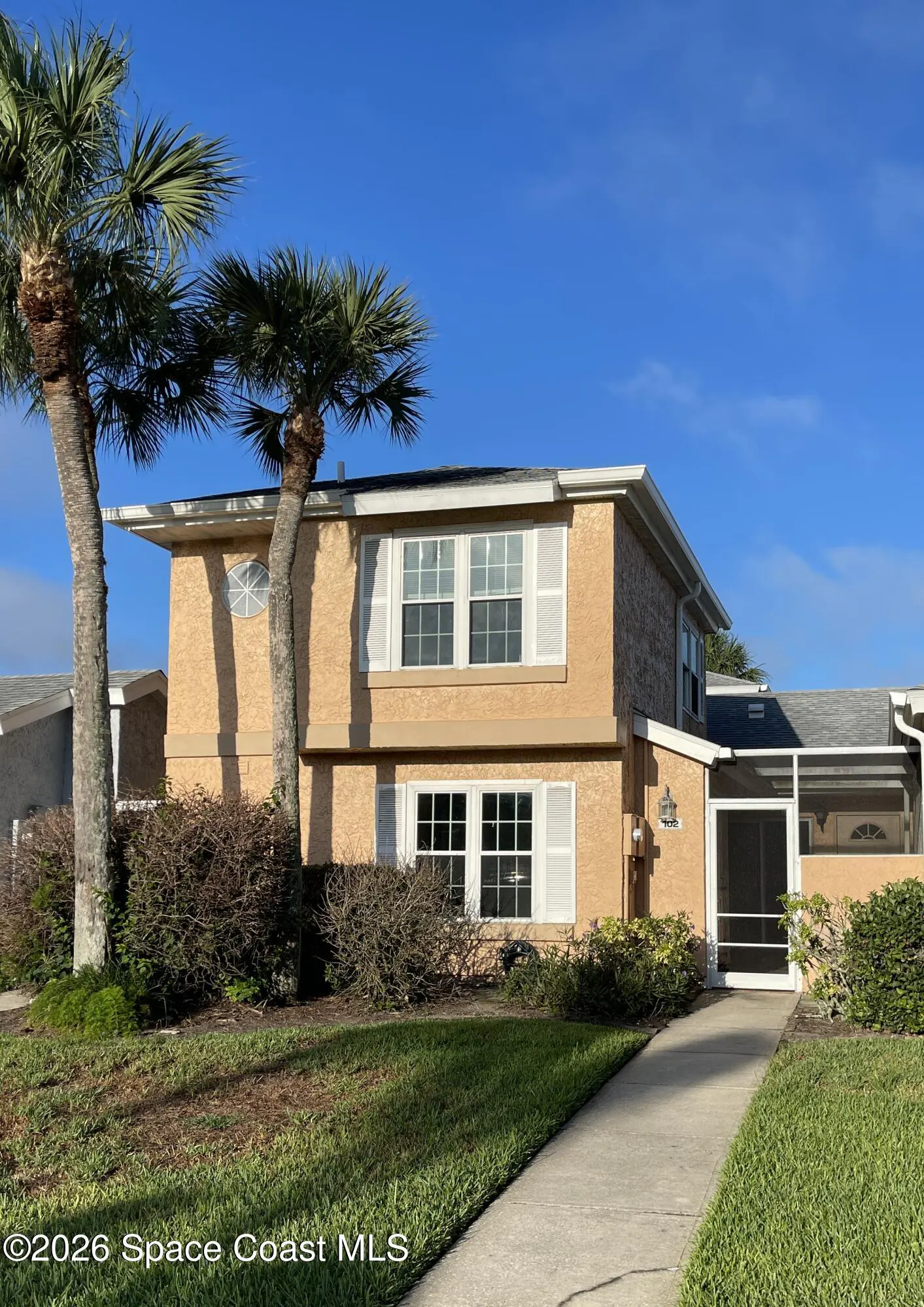 Two-story tan condo with white shutters and a screened-in porch. Palm trees flank the building under a clear blue sky.