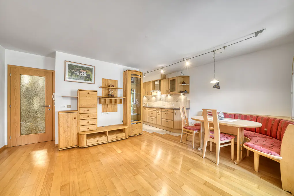 Bright, open-concept living space with light wood floors, cabinets, and furniture. A dining area with red-cushioned seating is visible.