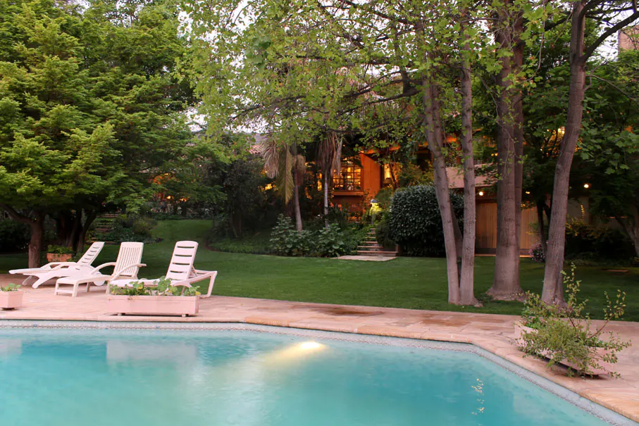 A backyard with a pool, lounge chairs, green grass, trees, and a house in the background.