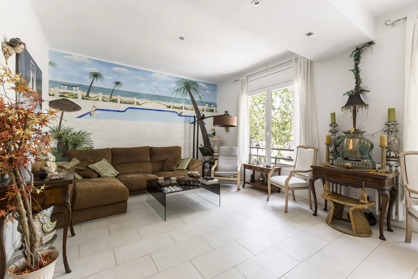 Bright living room with white tile floors, brown sectional sofa, and a mural of a beach scene with palm trees and a pool.