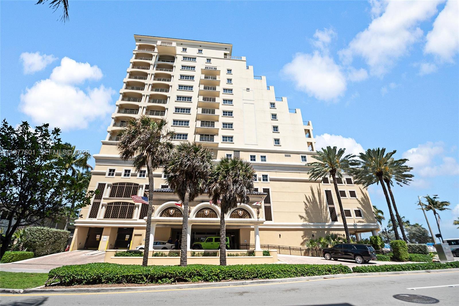 Low-angle view of a tall, beige tiered apartment building with palm trees, a green car, and a black SUV in front. [AI-generated text]