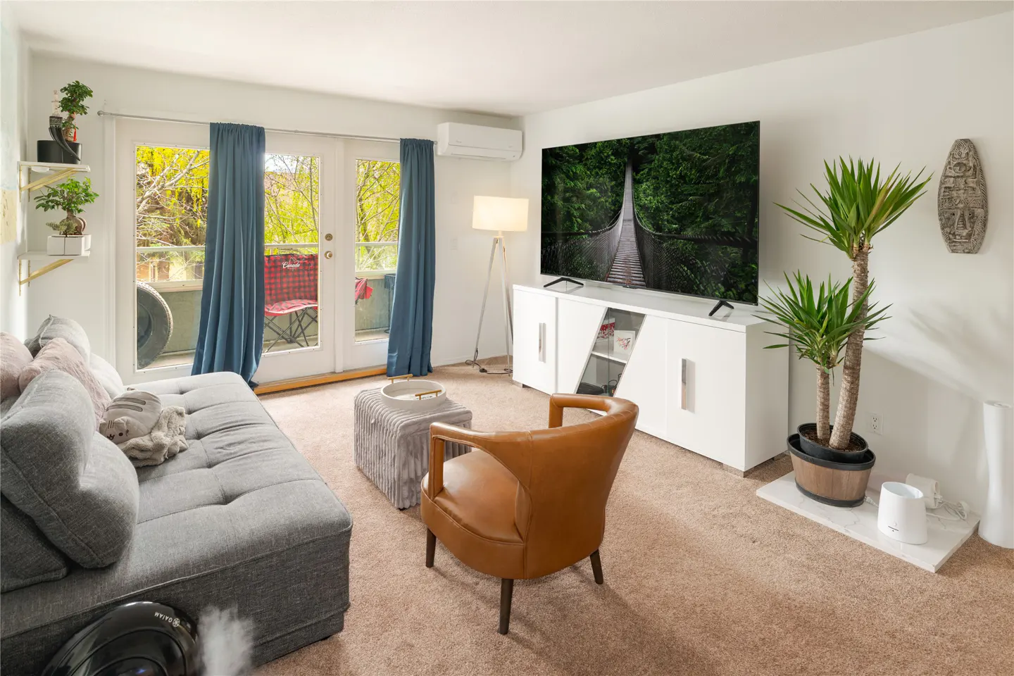 Living room with gray sofa, brown chair, TV showing a bridge, and balcony with blue curtains.
