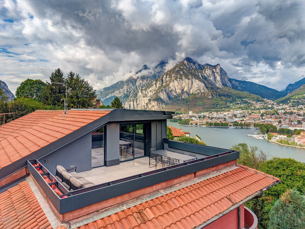 Modern rooftop terrace with outdoor seating overlooking a lake, town, and dramatic mountain range under a cloudy sky. [AI-generated text]