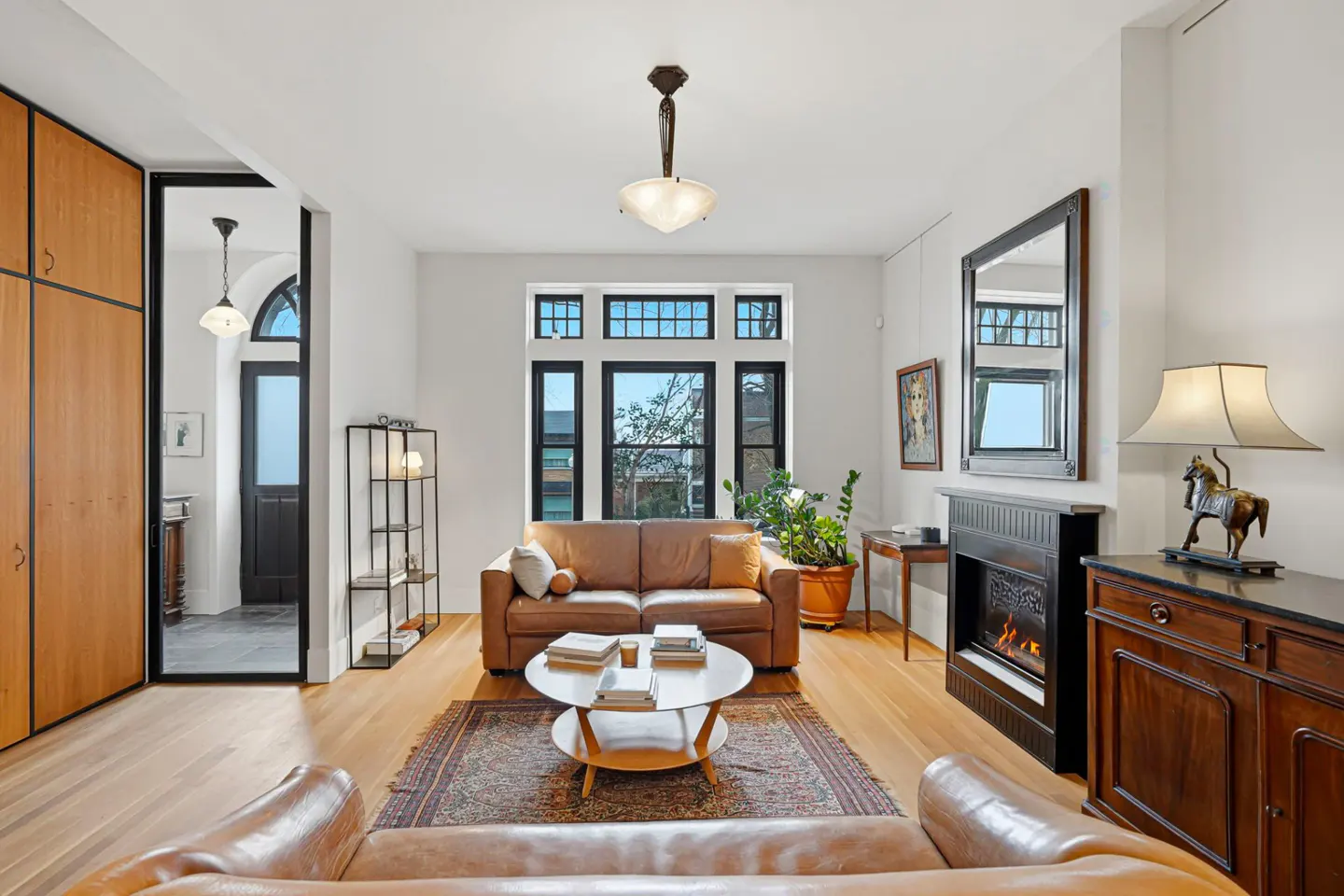Bright living room with hardwood floors, a brown leather sofa, a fireplace, and a large window. A rug sits under a round coffee table with books.
