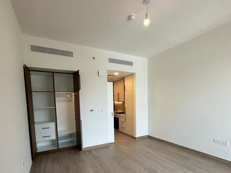 An apartment interior with wood floors, white walls, and an open closet. A kitchen is visible through a doorway.