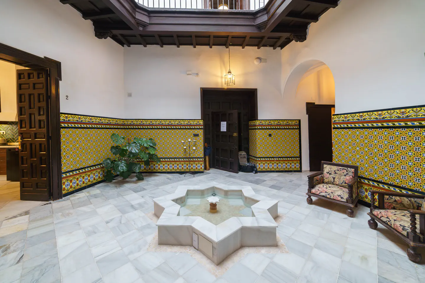 Interior courtyard with marble floors, yellow tiled walls, and a star-shaped fountain. Two chairs sit near a dark wooden door.