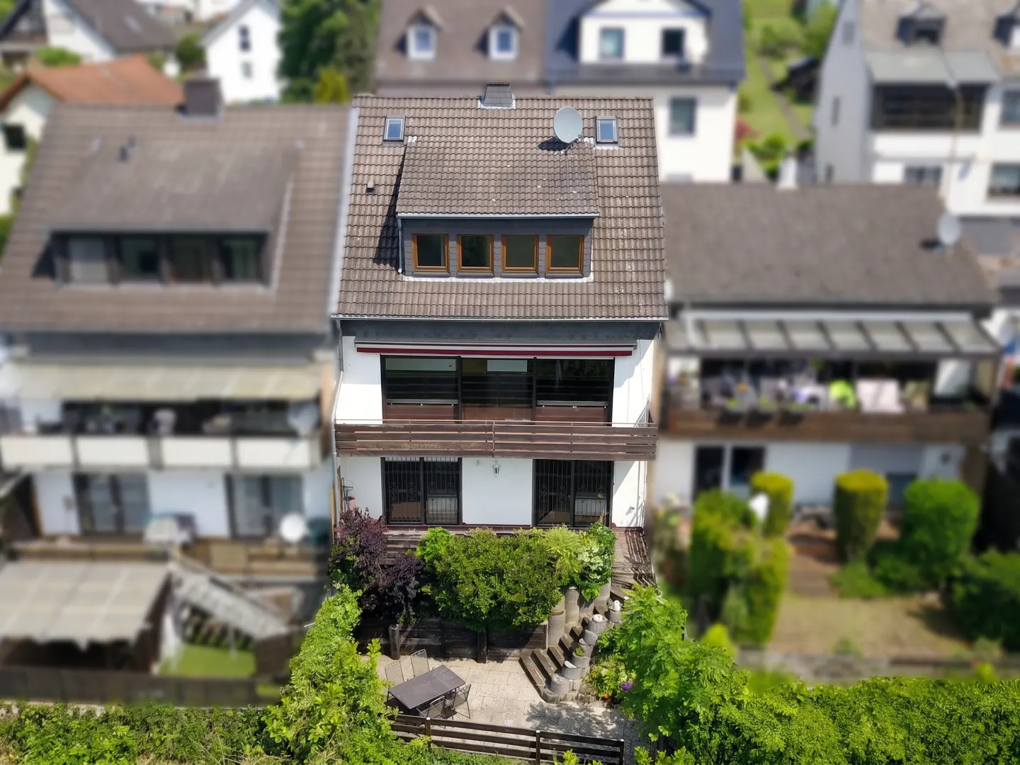 A three-story white house with a brown roof and a small garden. The house has a balcony and a patio with a table and chairs.