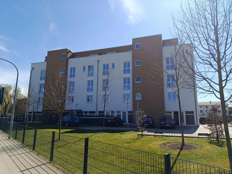 A modern, four-story apartment building with white and brown facade, cars parked in front, and a green lawn.