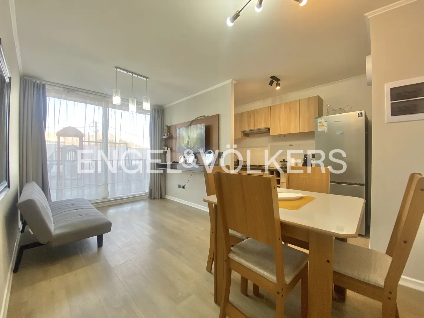 A bright apartment interior with a dining table, gray sofa, and kitchen with wooden cabinets and a stainless steel refrigerator.