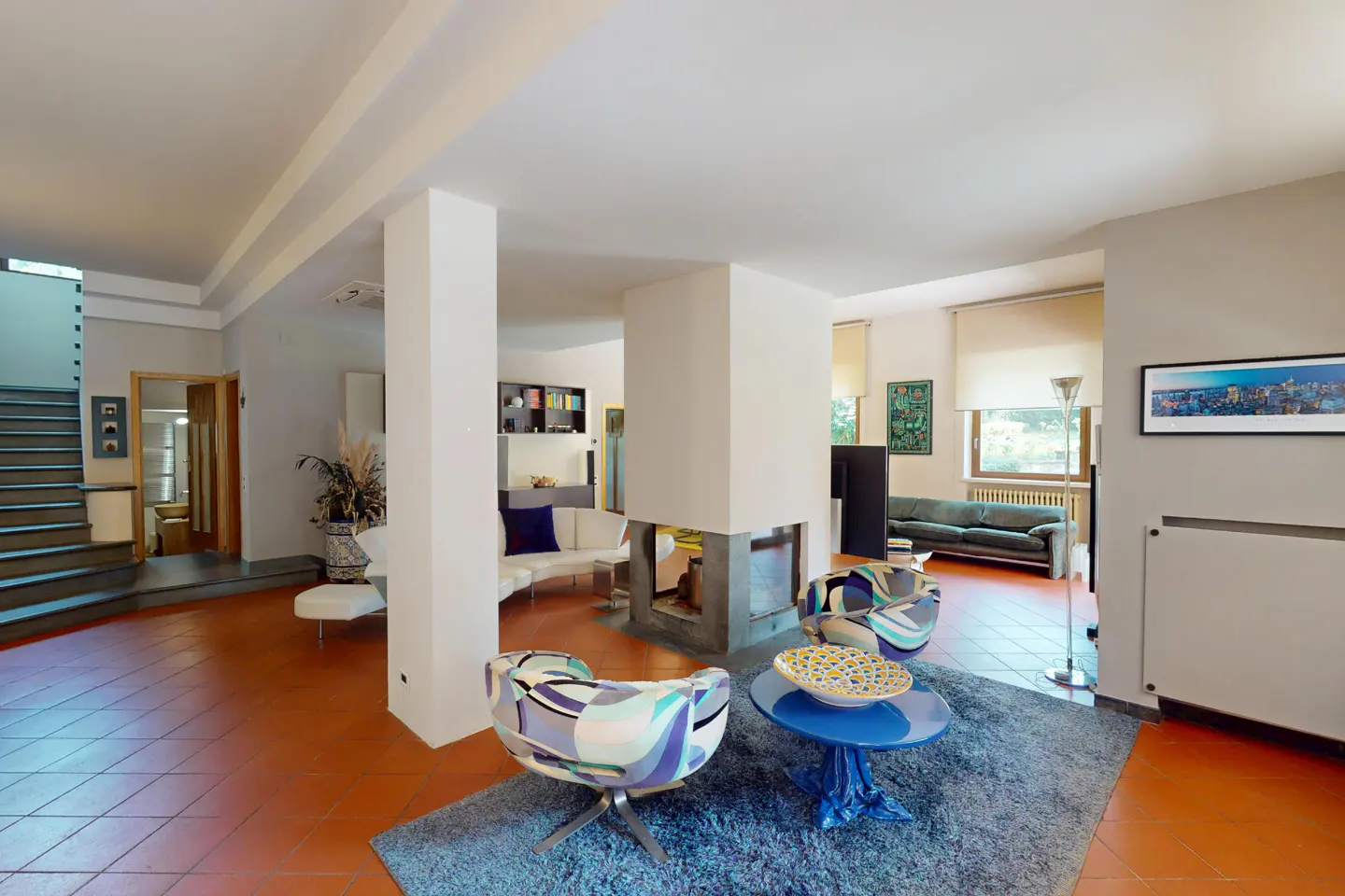 Open-concept living room with a fireplace, blue rug, and patterned chairs. A staircase and doorway are visible in the background.