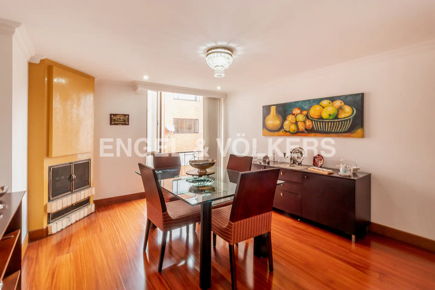 A dining room with a glass table, four chairs, a fireplace, and a painting of fruit.