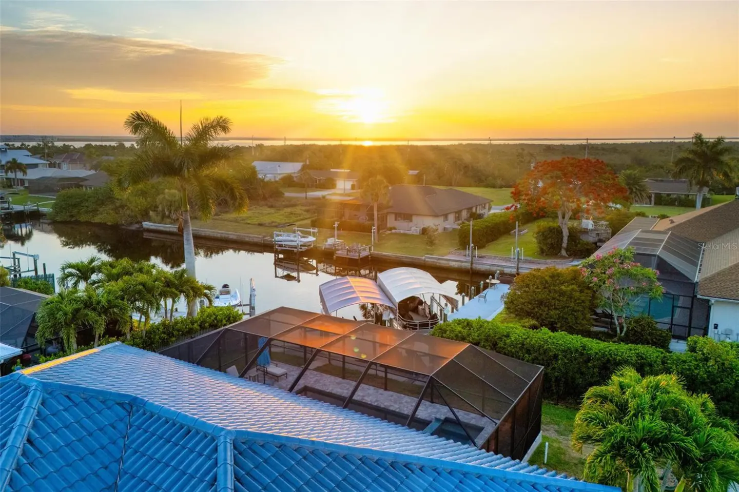 Aerial view of waterfront homes with boat docks on canals at sunset. Blue tiled roof in foreground.