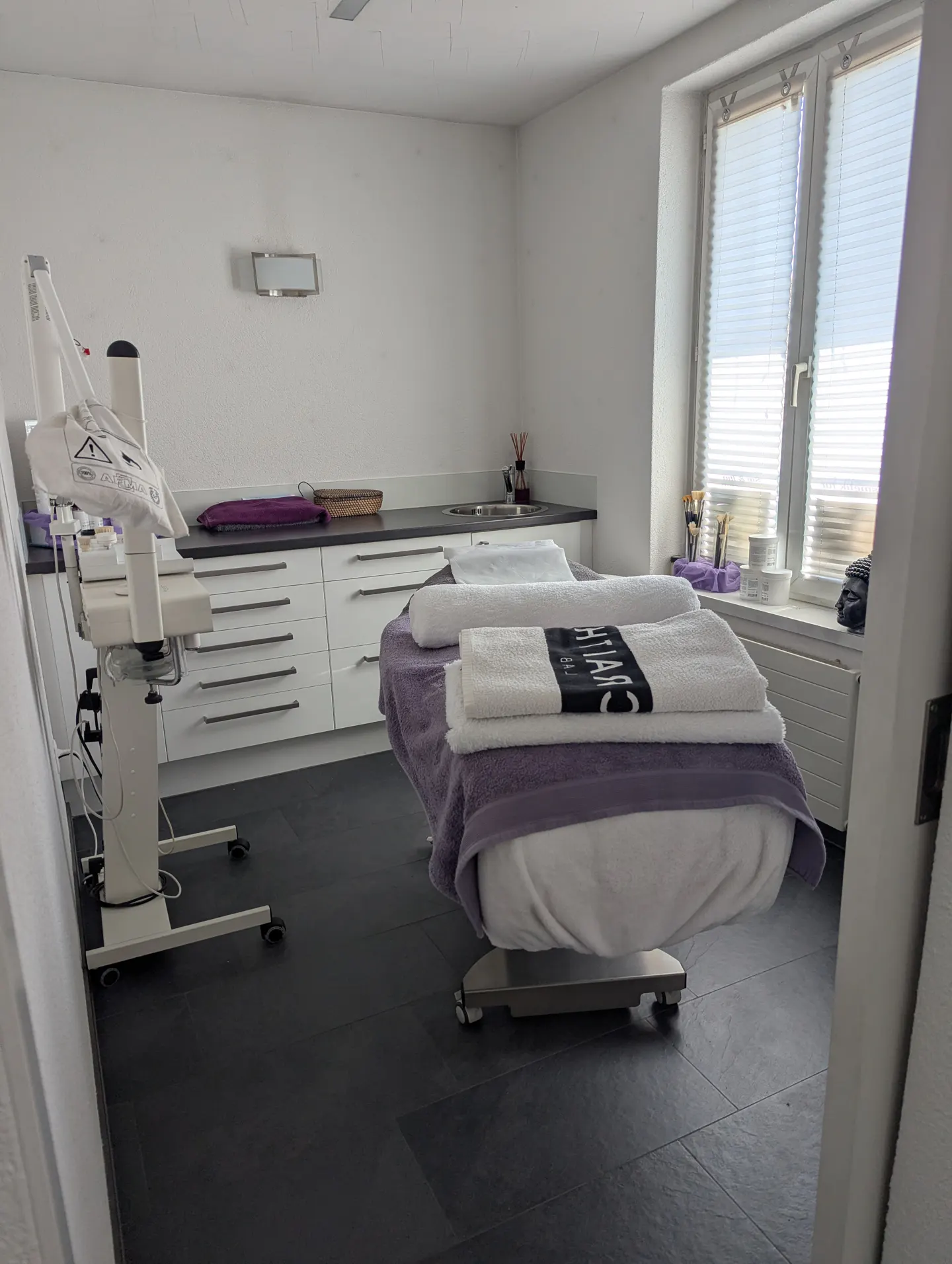 A bright treatment room with a bed covered in white towels and a purple blanket. A window with blinds is on the right.