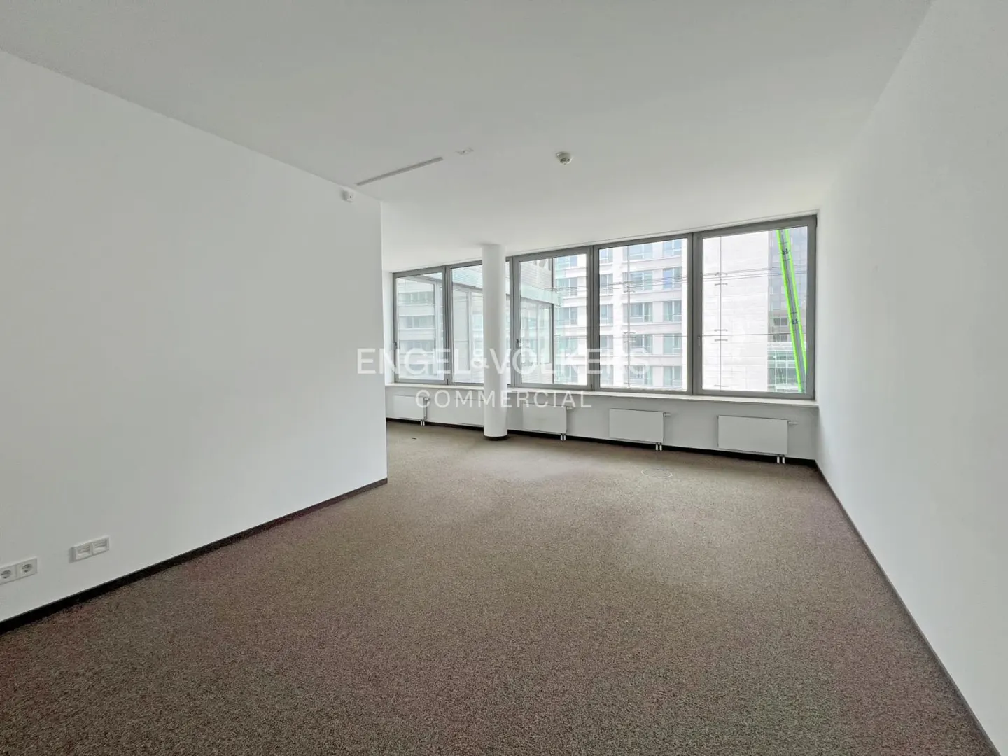 Bright, empty office space with brown carpet, white walls, and large windows overlooking a city. Radiators line the window wall.
