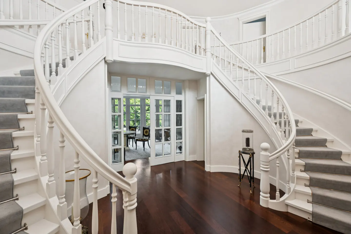 Elegant foyer with a winding white staircase and gray carpet runner. Dark wood floors lead to glass doors revealing a dining area.