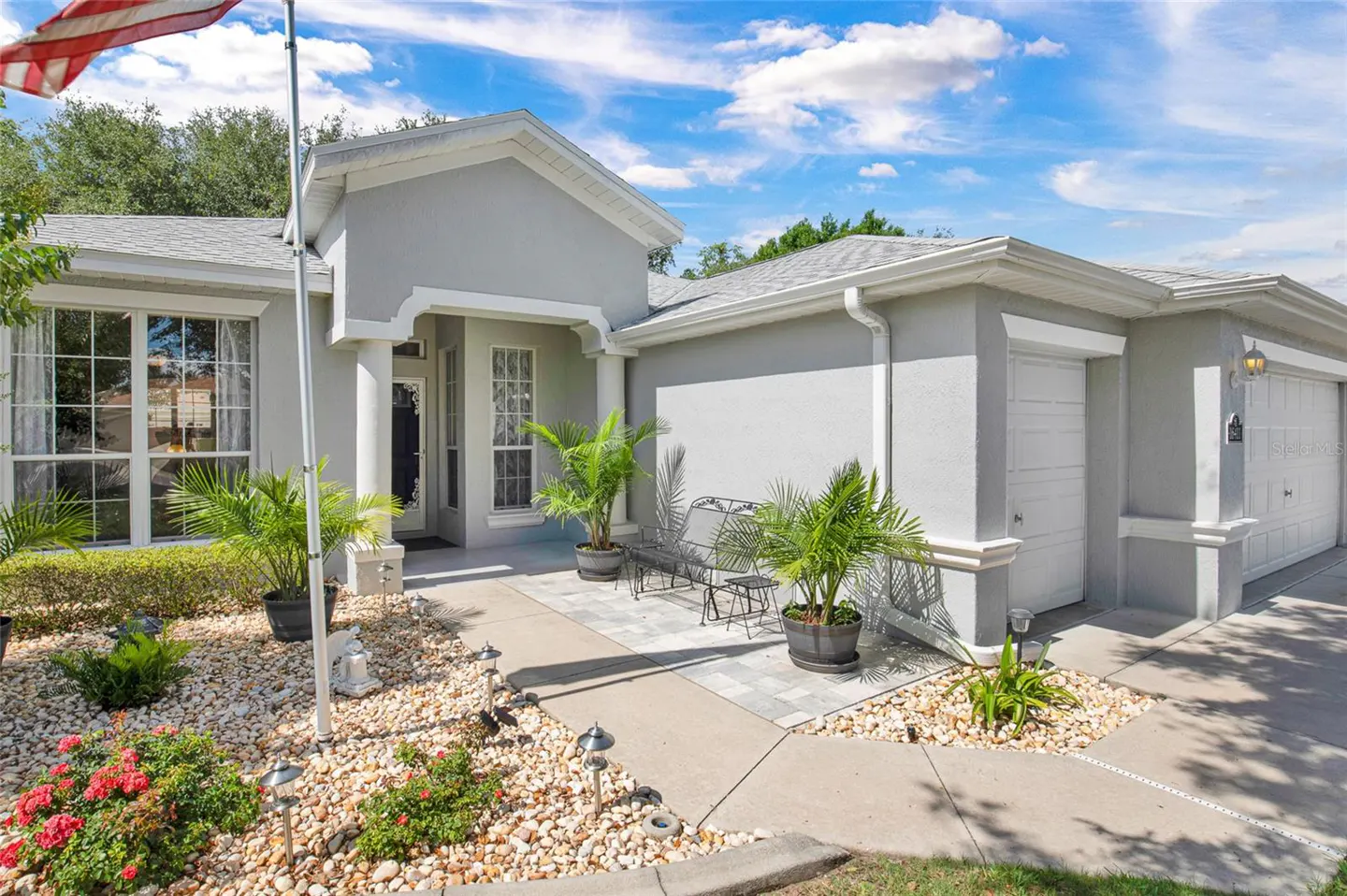 Single-story gray house with a white garage, stone landscaping, potted palms, and a small metal bench under a blue sky.