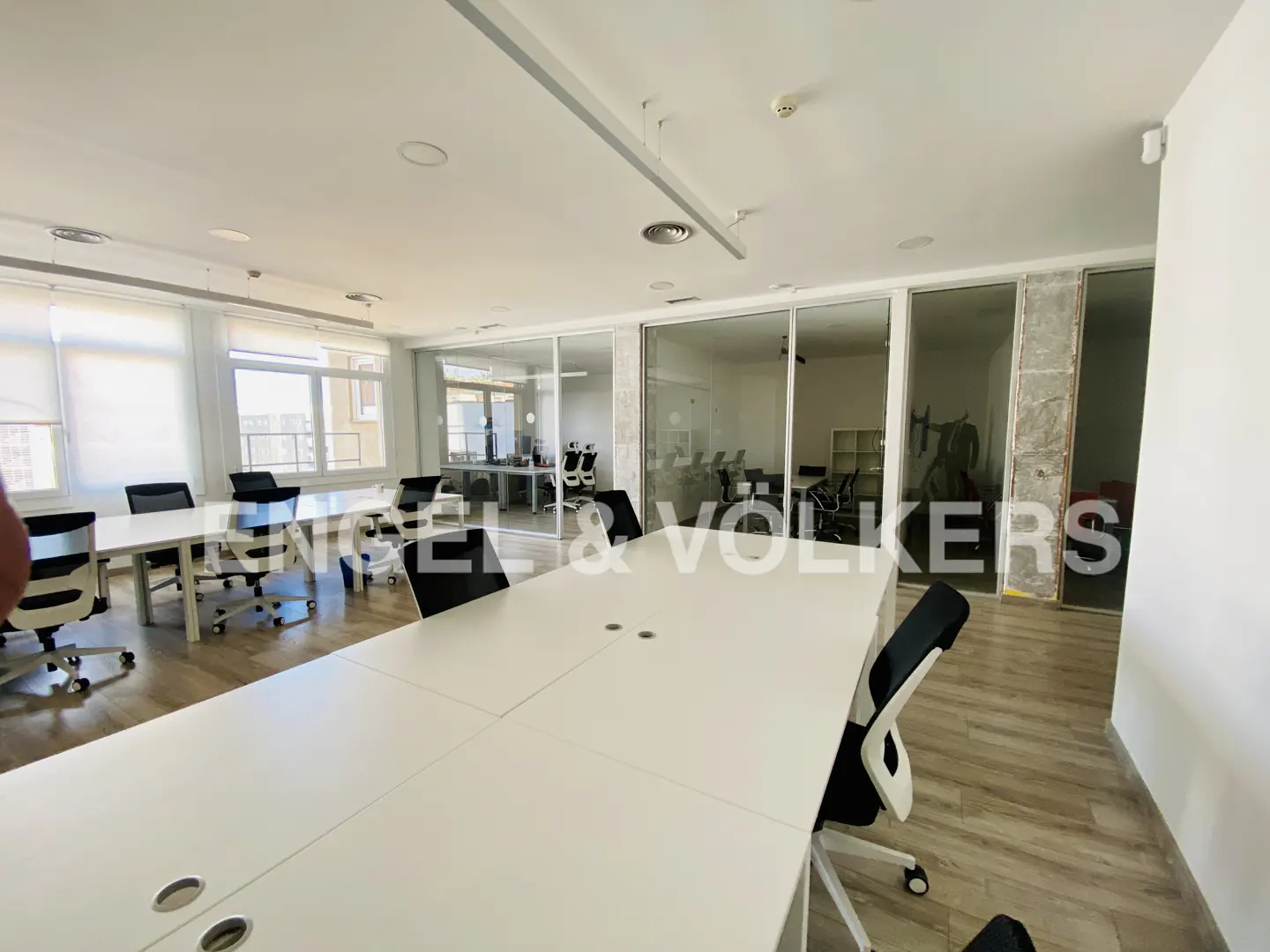 Bright office space with white tables, black chairs, and glass-walled rooms in the background.