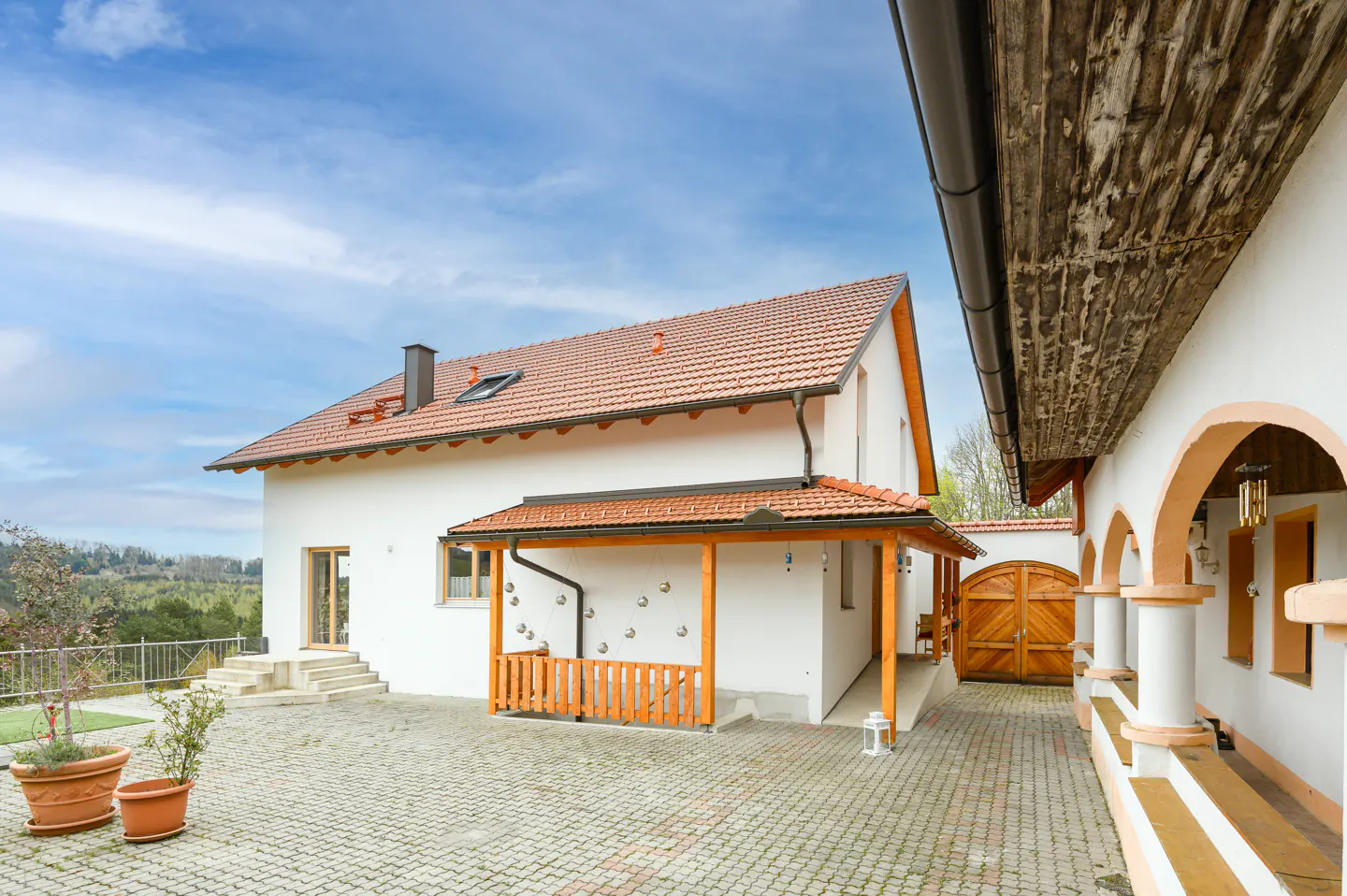 Exterior view of a white house with a red tile roof and wooden porch, set on a paved courtyard under a blue sky.