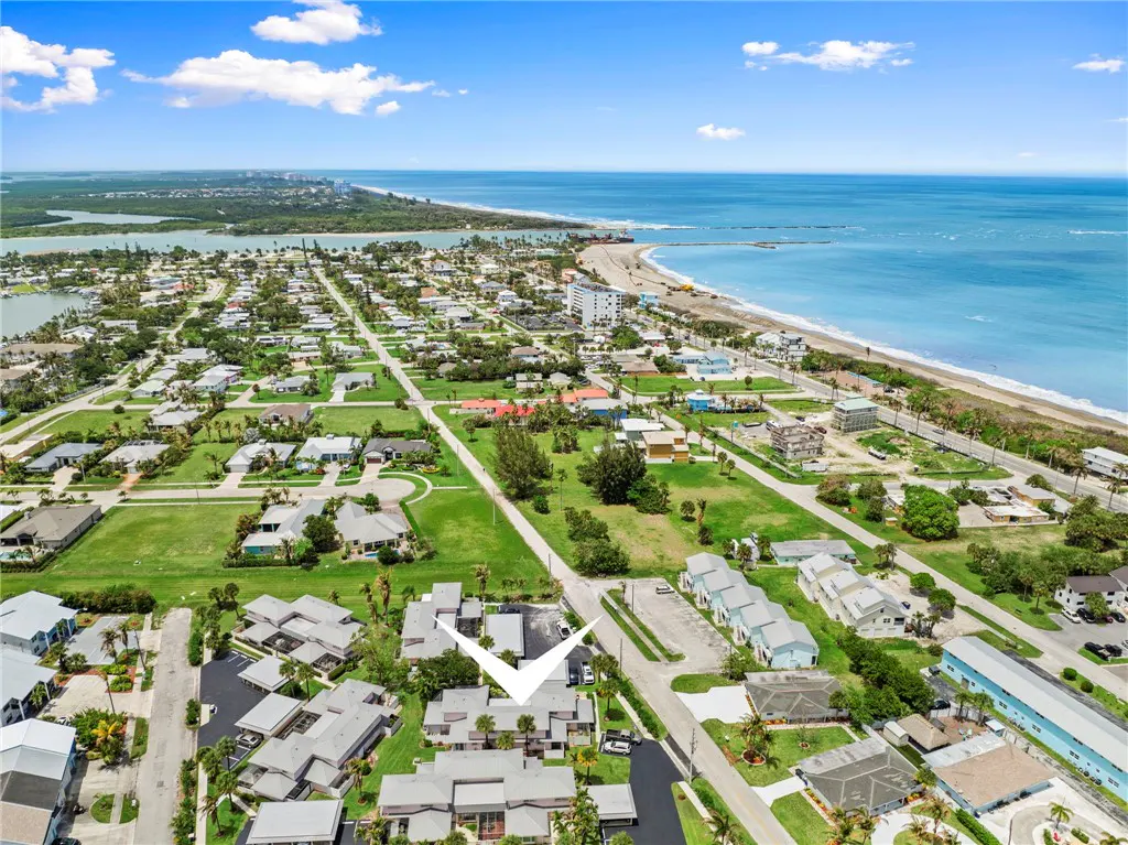 Aerial view of a coastal neighborhood with light gray condos, green lawns, and a white arrow pointing to a property near the beach.