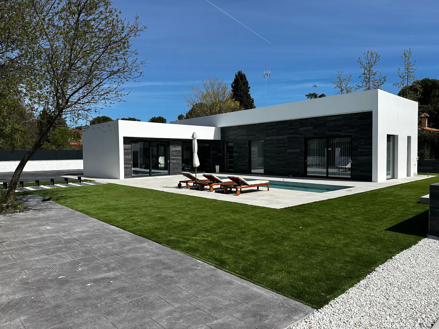 Modern, single-story home with white and dark stone exterior, pool, lounge chairs, and green lawn under a blue sky.