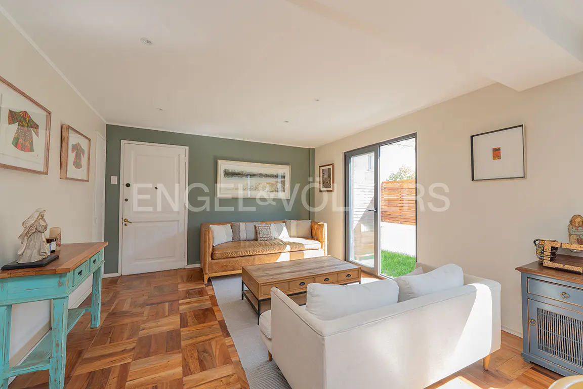 Living room with parquet floors, white sofa, and tan leather couch. A blue console table sits near framed art. Sliding glass doors lead to a green lawn.