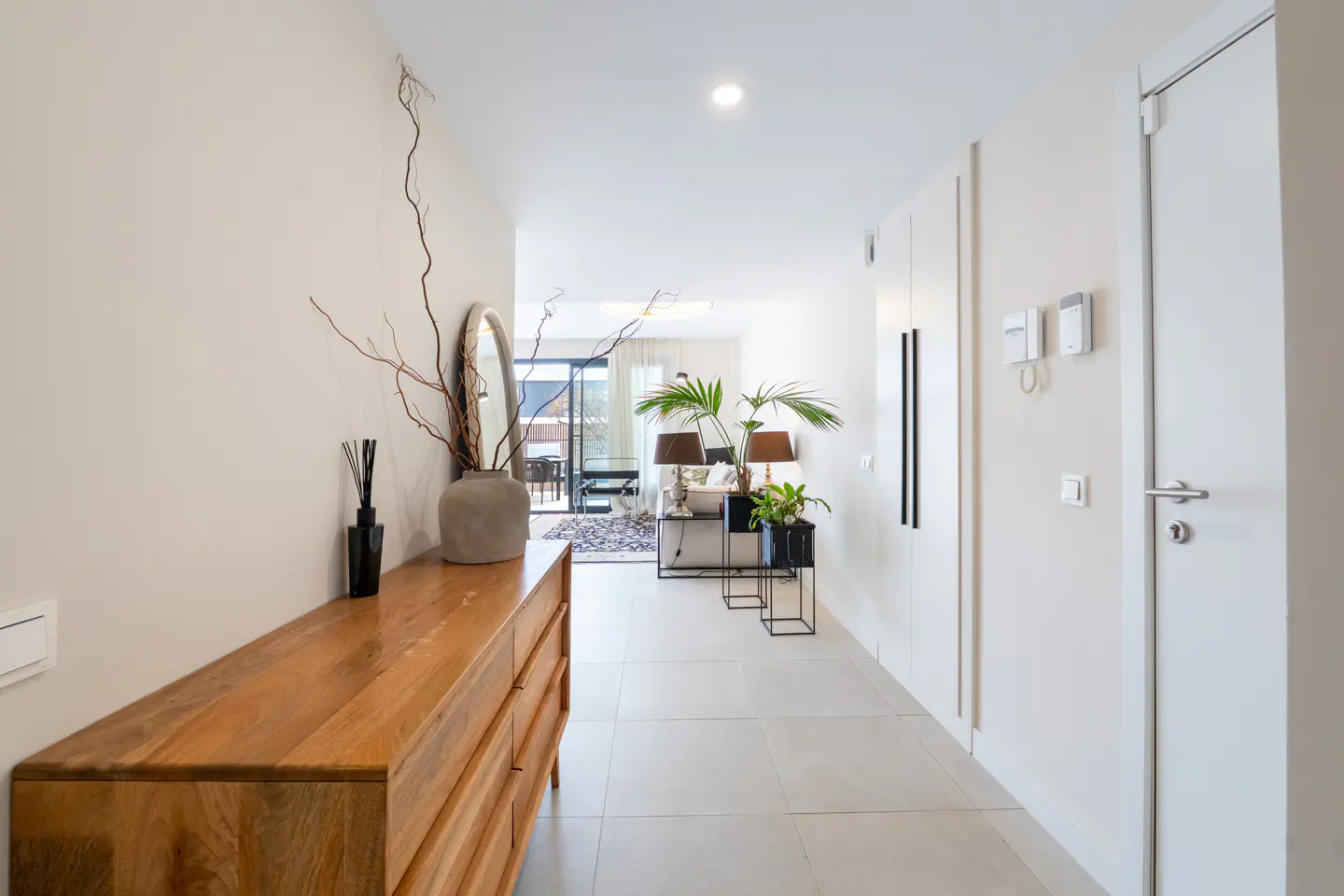 Bright hallway with white walls and tile floor. A wooden dresser sits to the left with a mirror and vase. Living room visible at the end.
