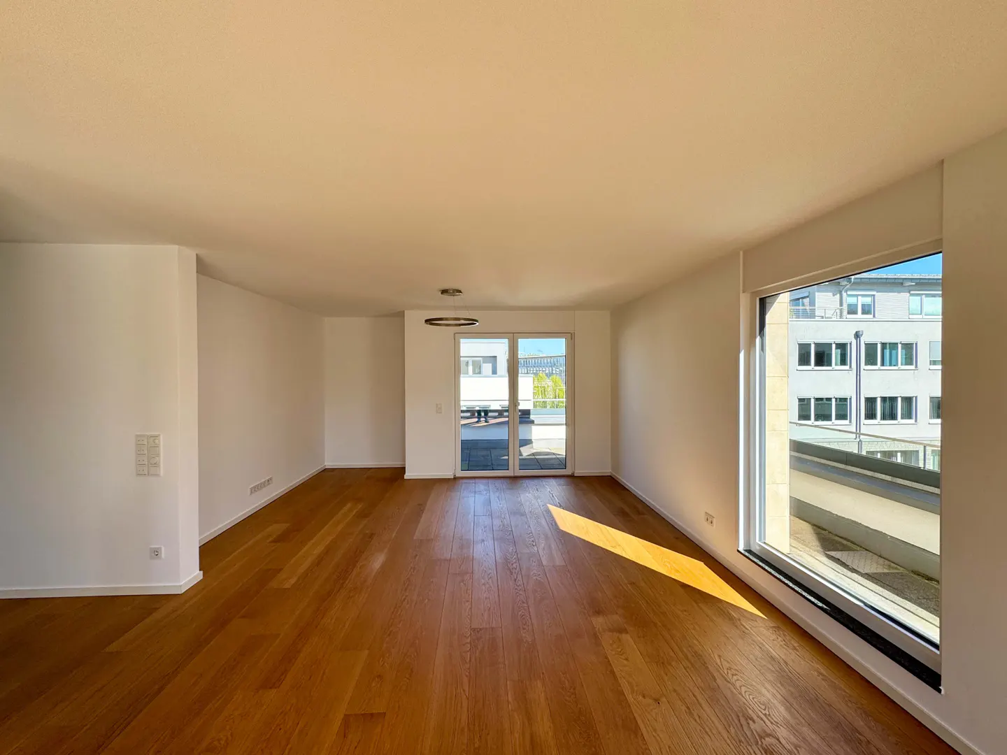 Bright, empty room with hardwood floors, white walls, and a large window. Sunlight streams across the floor. Glass doors lead to a balcony.