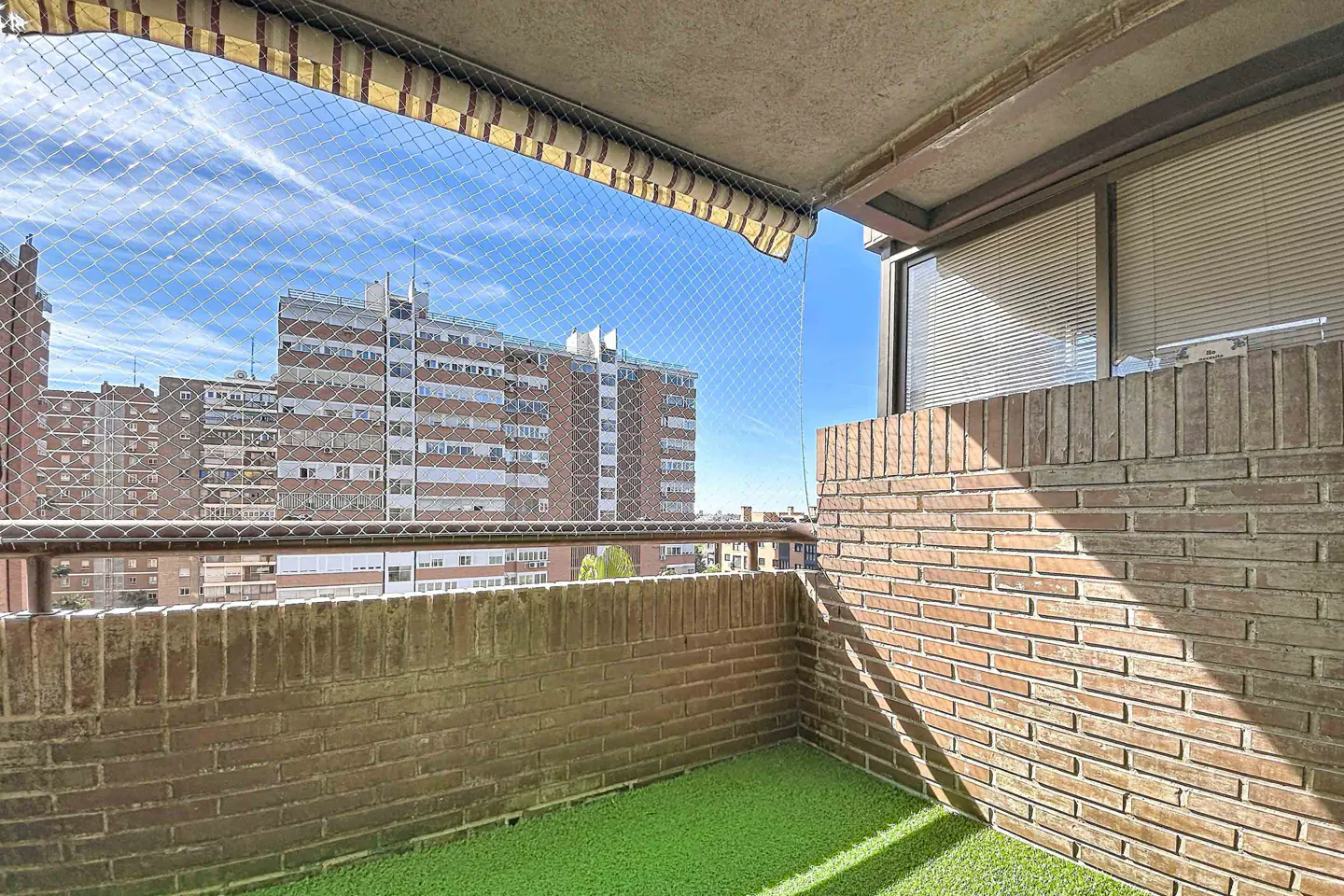 Balcony view with brick walls, green turf, and safety netting. Buildings are visible in the background under a blue sky.