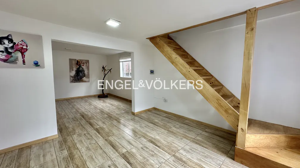 A bright room with wood-look tile floors, white walls, and a wooden staircase. Artwork hangs on the walls. The Engel & Völkers logo is superimposed.
