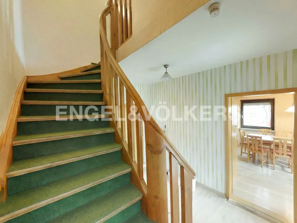 Interior view of a home featuring a wooden spiral staircase with green carpet, striped wallpaper, and a dining room visible through a doorway.