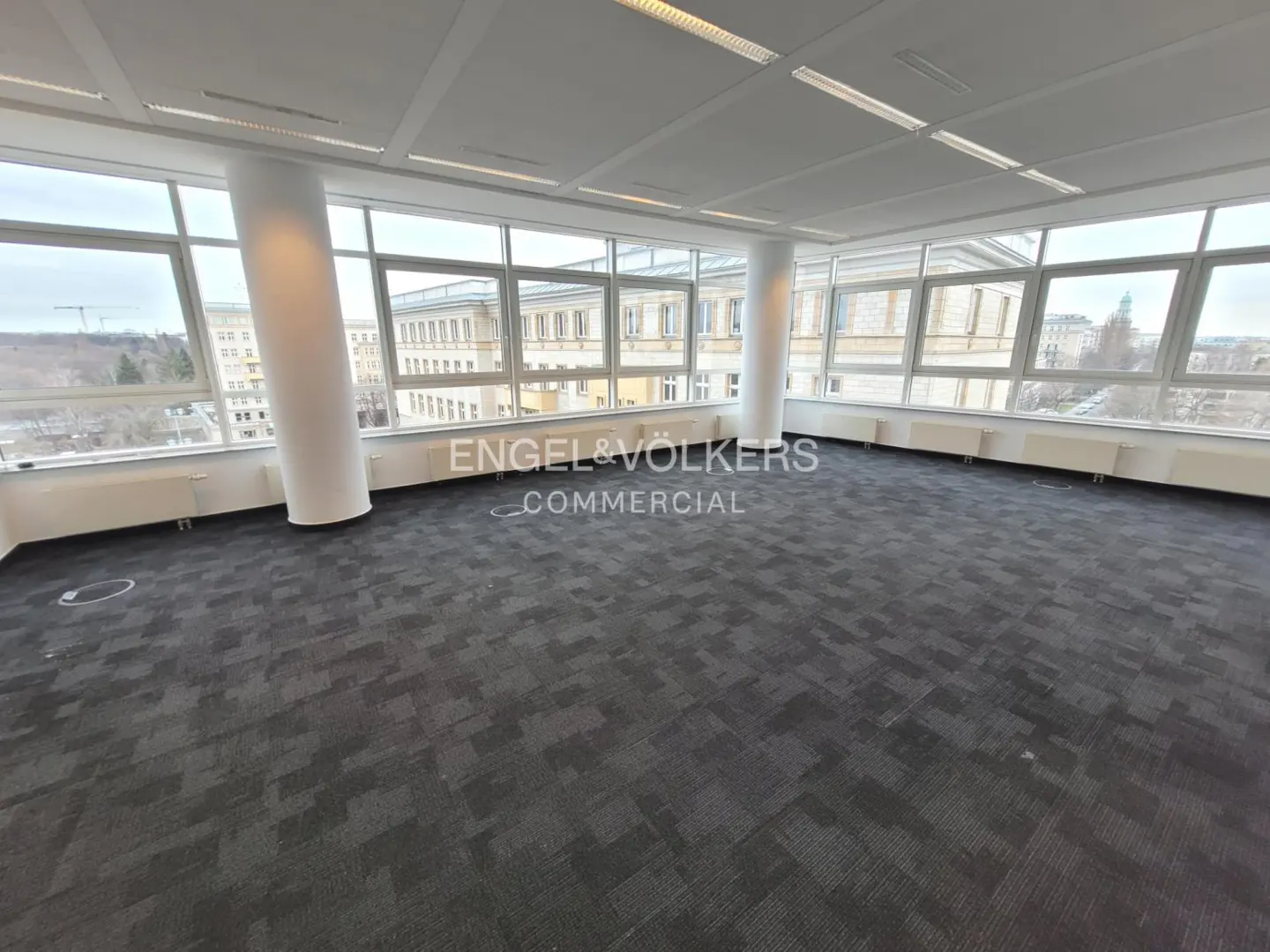 Bright, empty office space with gray carpet, white walls, and large windows overlooking a city. Engel & Völkers Commercial logo visible.