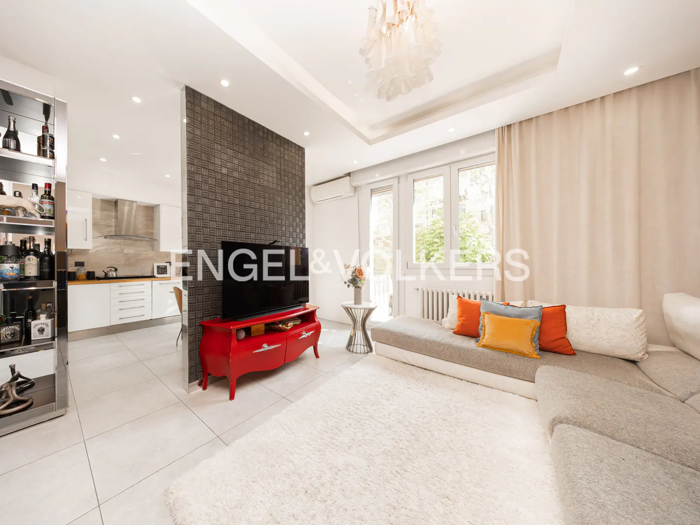 Bright living room with a gray sectional sofa, orange pillows, and a red TV stand. A modern kitchen is visible in the background.
