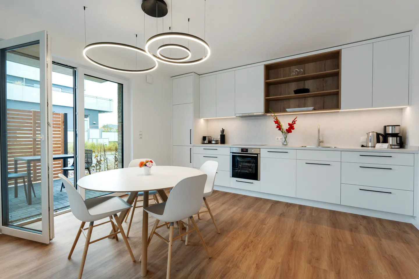 Bright kitchen with white cabinets, wood floor, and round table with four chairs. Open patio door to outdoor seating.