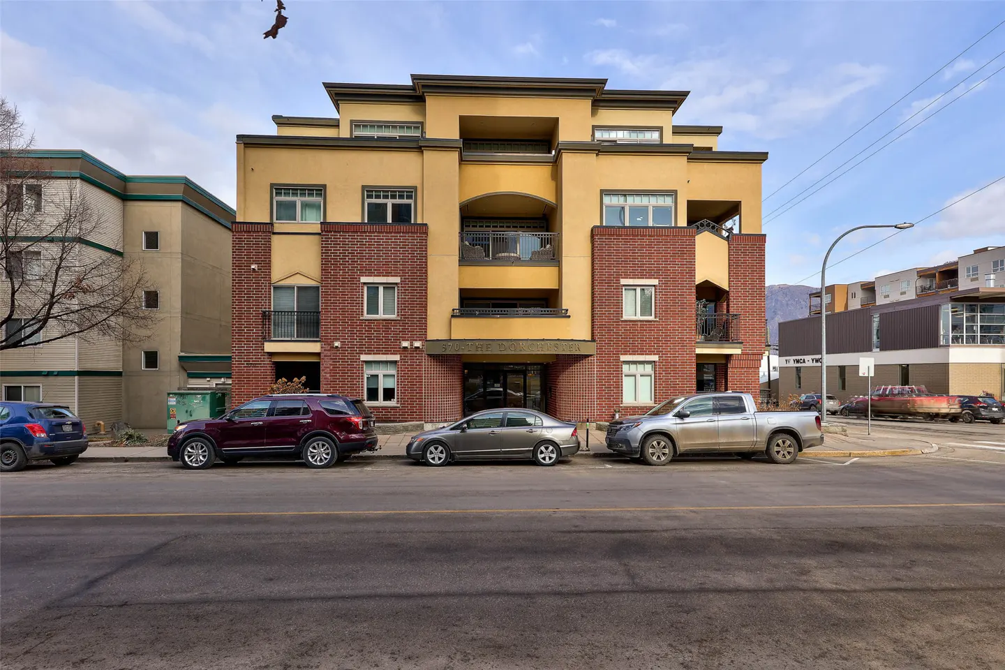 A four-story apartment building with yellow stucco and red brick accents. Cars are parked along the street in front.