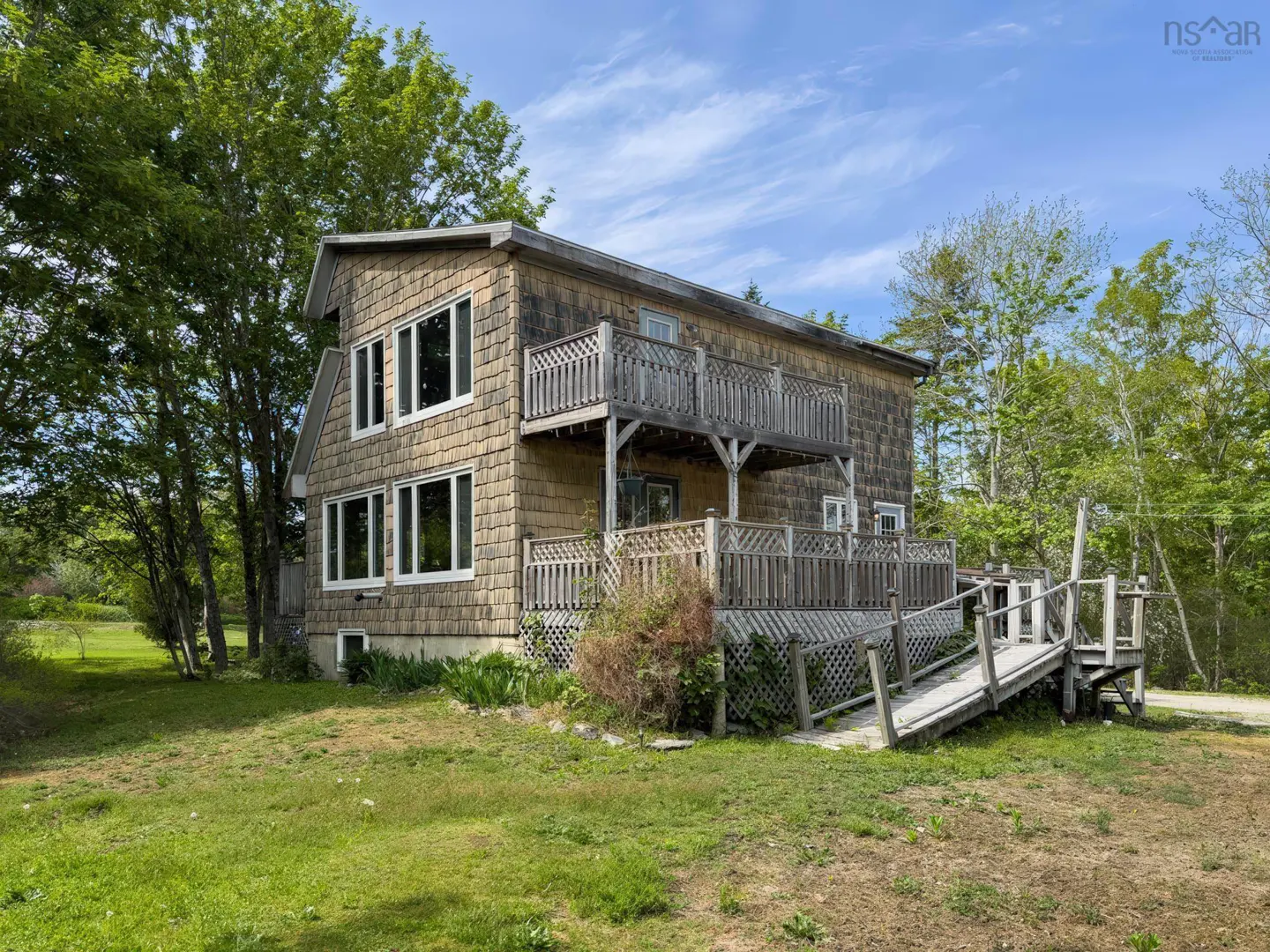 Two-story house with cedar shingles, a wooden deck, a balcony, and a wheelchair ramp, surrounded by trees on a sunny day.